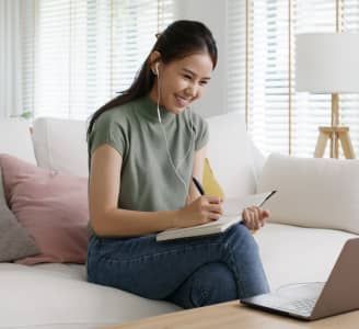 Smiling woman studying online at home, wearing earphones and taking notes in a notebook while looking at a laptop